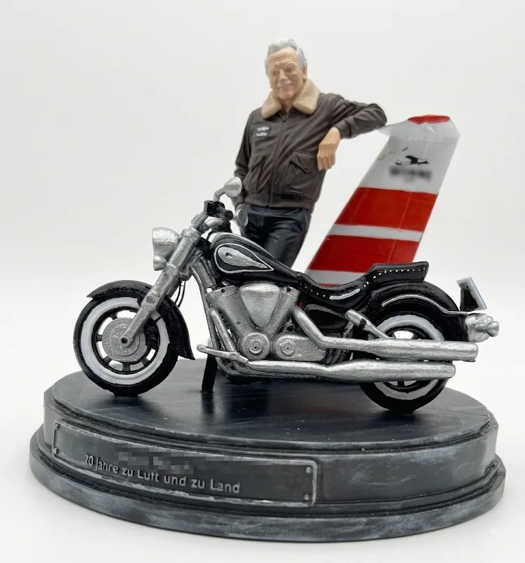 Personalisierte Figur zum 70. Geburtstag: Mann in Fliegerjacke am Heckfl&uuml;gel eines Flugzeugs, daneben ein nachgebautes Motorrad auf einem Sockel mit Widmung